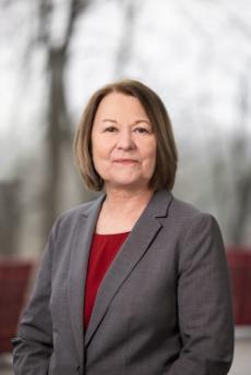 A headshot of a woman in a gray blazer with a red shirt.